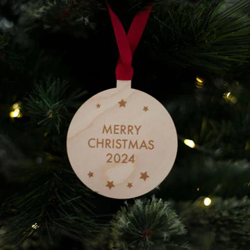 A wooden christmas decoration on a christmas tree which says Merry Christmas 2024 in braille, tied with a red ribbon 